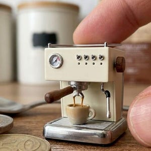 May include: A miniature cream-colored espresso machine with a silver pressure gauge, four small toggle switches, a wooden portafilter handle, and a steam wand. A tiny ceramic cup sits on the drip tray, filled with a dark brown liquid coffee.