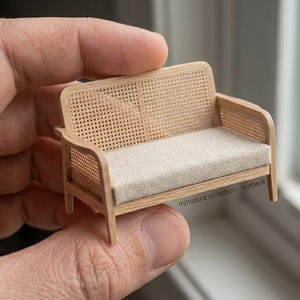 Pode incluir: Um sofá de dois lugares em miniatura com estrutura de madeira clara, painéis de palhinha trançada nas costas e laterais, e uma almofada de assento em tecido bege. A pequena peça de mobiliário inclui o texto miniature collection - loveseat.