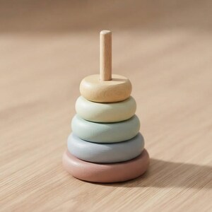 May include: A wooden stacking toy featuring a central vertical post and five smooth, rounded rings in graduated sizes. The rings are painted in soft, muted pastel colors including yellow, light green, sage, blue-gray, and a dusty rose pink base.