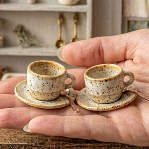 以下が含まれることがあります： クリーム色とタン色の斑点模様の釉薬が施された、陶器製のミニチュアコーヒーカップ2客とソーサーのセット。各カップにはソーサーの上に小さな金属製のスプーンが添えられており、ドールハウス用の手作りデザインとなっています。