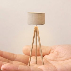 May include: A miniature floor lamp featuring a light-colored wooden tripod base and a cylindrical beige fabric lampshade. The lamp is constructed with three thin legs that meet at a central point beneath the shade, creating a compact design.