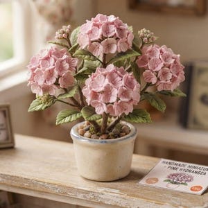 May include: A miniature potted plant featuring three clusters of light pink hydrangea flowers with green leaves, set in a small, off-white ceramic pot filled with pebbles. A small card next to the pot reads HANDMADE MINIATURES - PINK HYDRANGEA.