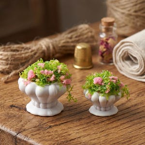 Puede incluir: Dos pequeñas urnas decorativas blancas llenas de arreglos florales verdes y rosas. Las urnas están sobre una superficie de madera, con un dedal, hilo y un pequeño frasco de vidrio con un tapón de corcho en el fondo.