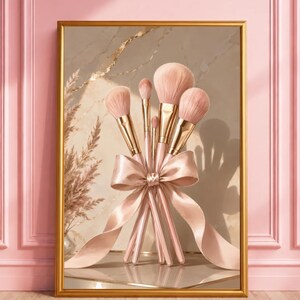 Könnte beinhalten: Ein gerahmter Wandkunstdruck mit einer Sammlung von fünf Make-up-Pinseln mit metallisch goldenen Zwingen und weichen rosa Borsten, die mit einem großen, eleganten Satinband in einem passenden hellen Rosaton vor einem neutralen Hintergrund gebunden sind.