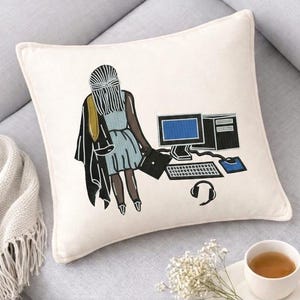 Può includere: Un cuscino quadrato color bianco sporco con un'illustrazione ricamata di una persona con lunghi capelli scuri che indossa un vestito azzurro e una giacca nera, in piedi vicino a una postazione computer con monitor, tastiera, mouse e cuffie.