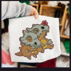 Puede incluir: Una sudadera blanca con un diseño bordado de dos criaturas marrones con cascos grises en forma de cráneo. Una criatura sostiene un hueso, mientras que la otra tiene una pequeña flor roja en la cabeza. El bordado utiliza hilo marrón, gris y rojo.
