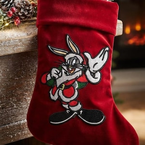 May include: A red velvet Christmas stocking featuring an embroidered patch of Bugs Bunny wearing a Santa Claus suit. The character is depicted with long ears, white gloves, and black shoes, centered on the front of the plush red fabric stocking.