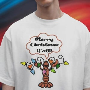 Könnte beinhalten: Ein weißes T-Shirt mit einer Stickerei, die einen Hummer zeigt, der sich in bunten Weihnachtsbeleuchtungen verfangen hat. Über dem Hummer befindet sich eine Sprechblase mit dem schwarz gestickten Text Merry Christmas Y'all! Das Design verwendet Orange, Blau, Grün und Pink.