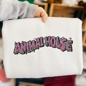 May include: A white crewneck sweatshirt featuring the text ANIMAL HOUSE embroidered in a stylized, arched font. The letters are filled with a textured pink thread and outlined in black and light blue, creating a bold, graphic appearance.