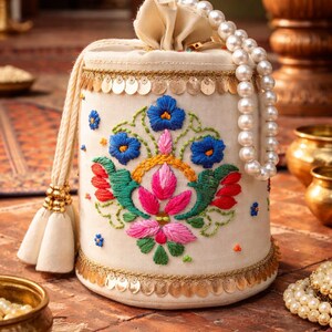 Könnte beinhalten: Cremefarbener, bestickter Beutel mit Kordelzug und einem floralen Design in Blau, Rosa, Grün und Gold. Die Tasche ist mit goldenen Pailletten und einer cremefarbenen Quaste verziert. Eine Perlenkette hängt von oben.