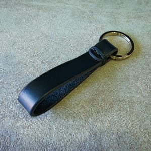 May include: A black leather keyring featuring a smooth, folded strap design secured with a matching leather loop and a circular silver-toned metal split ring for attaching keys. The leather has a matte finish on the exterior and a textured underside.