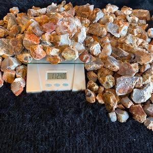 May include: A large collection of raw, unpolished agate stones in shades of brown, tan, gray, and orange. A white digital kitchen scale is positioned beneath a portion of the rocks, displaying a weight reading of 12.40 ounces on its screen.
