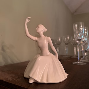 Può includere: Una statuetta in ceramica color bianco sporco dalla superficie liscia che raffigura una ballerina seduta con un braccio sollevato con grazia sopra la testa. La figura presenta una gonna semplice e fluida con pieghe sottili ed elegante.