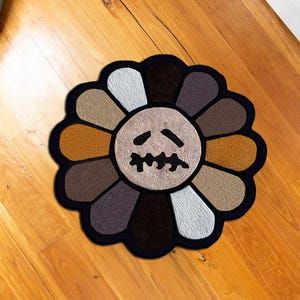 May include: A round, flower-shaped rug with a black border. The petals are various shades of brown, tan, and white, surrounding a beige center with a stylized, sad face. The rug is placed on a wooden floor.