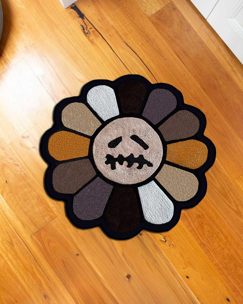 May include: A round, flower-shaped rug with a black border. The petals are various shades of brown, tan, and white, surrounding a beige center with a stylized, sad face. The rug is placed on a wooden floor.