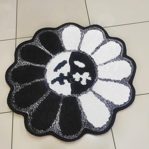 May include: A black and white flower-shaped rug. The rug features a design with alternating black and white petals. The center of the flower has a face design. The rug is placed on a tiled floor.