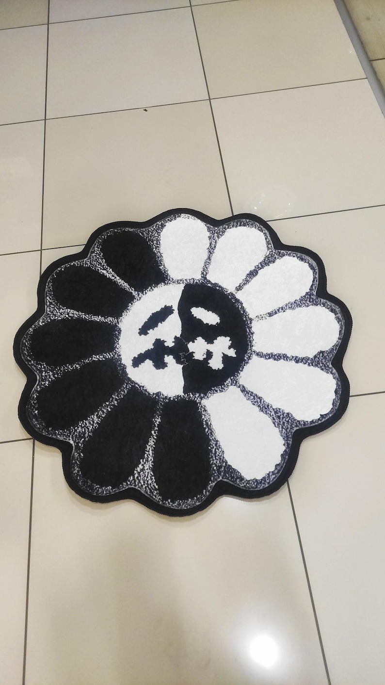 May include: A black and white flower-shaped rug. The rug features a design with alternating black and white petals. The center of the flower has a face design. The rug is placed on a tiled floor.