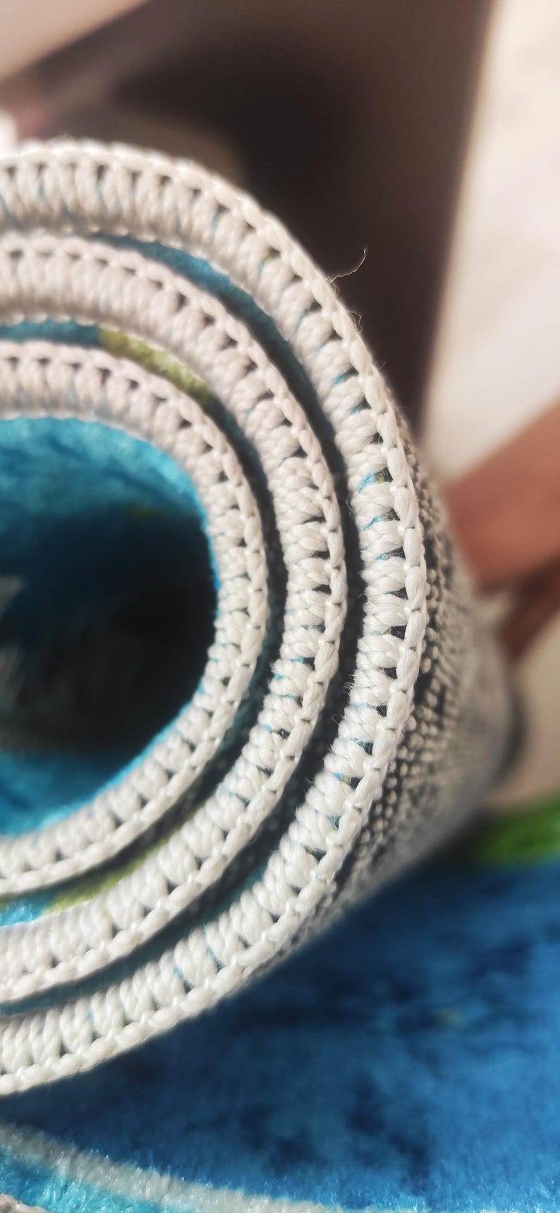 May include: Close-up of a rolled-up rug with a white, textured border. The rug's interior features a vibrant blue and green pattern. The white border is made of a thick, woven material.