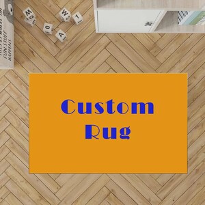 May include: A rectangular rug with a solid orange background and the text Custom Rug printed in blue letters in the centre. The rug is positioned on a light brown wooden floor with a herringbone pattern, near white wooden blocks and furniture.