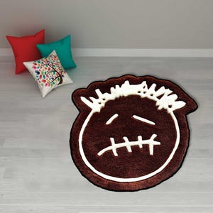 May include: A brown rug with a white outline and a cartoon face design. The rug has a stitched mouth and two horizontal lines for eyes. The rug is on a light wood floor with three pillows in the background.