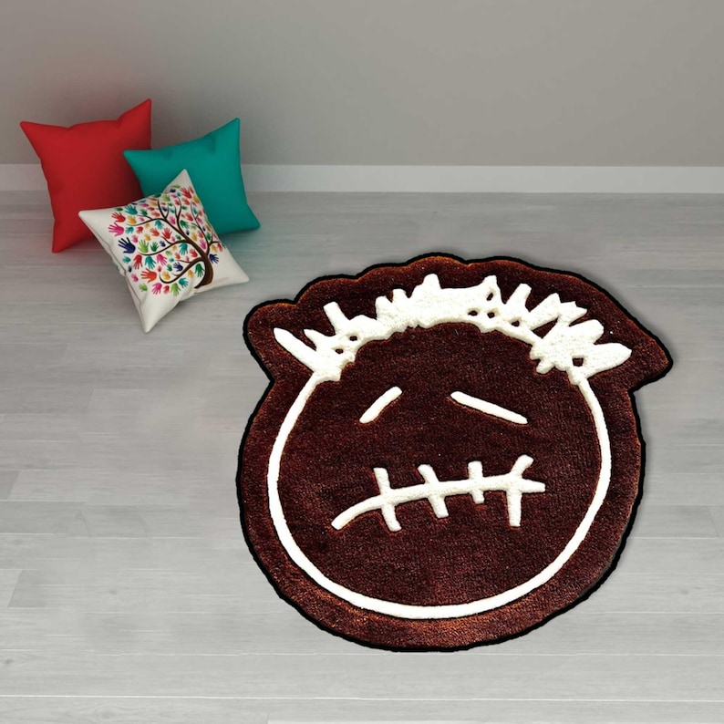 May include: A brown rug with a white outline and a cartoon face design. The rug has a stitched mouth and two horizontal lines for eyes. The rug is on a light wood floor with three pillows in the background.