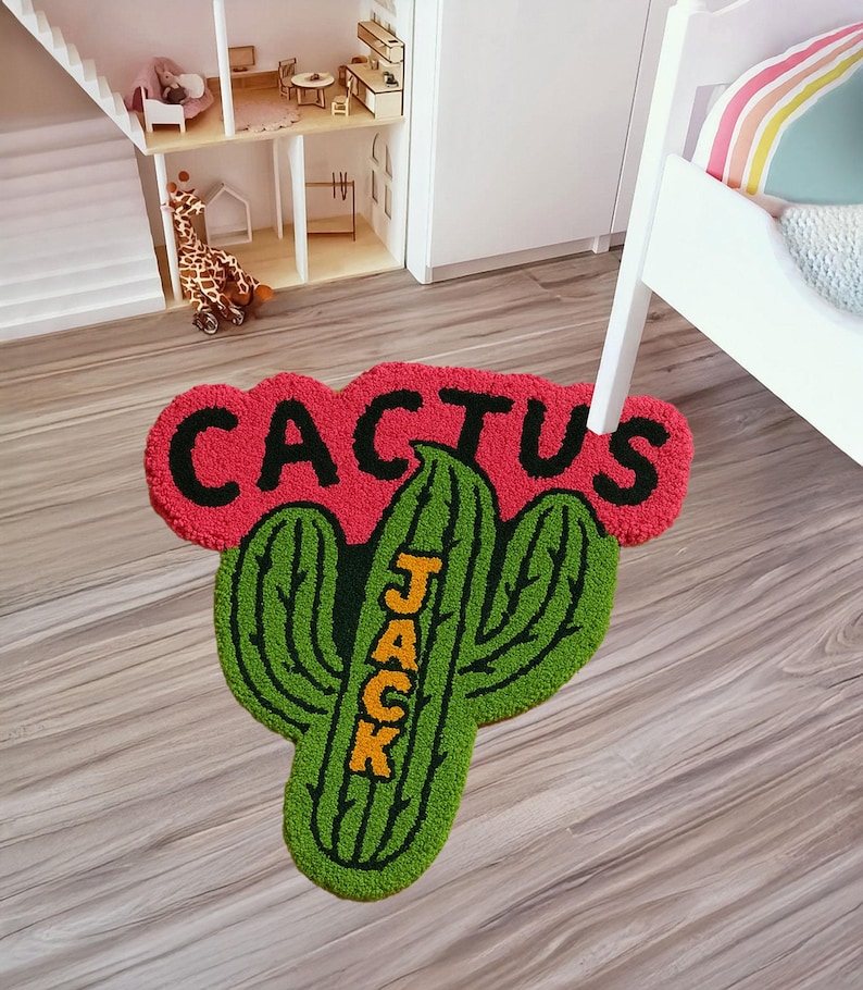 May include: A green cactus-shaped rug with the name "JACK" in orange letters on the cactus and the word "CACTUS" in black letters on a red background. The rug is on a wooden floor.