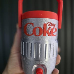May include: A silver and red Diet Coke water cooler with a red handle and a red tap. The Diet Coke logo is in red on the front. The cooler has a textured surface and is made of plastic. It is designed to hold and dispense cold drinks.