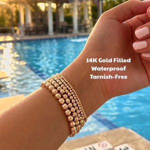 May include: A gold beaded bracelet is worn on a wrist. The bracelet is made of small, round beads. Text on the image reads: "14K Gold Filled Waterproof Tarnish-Free."
