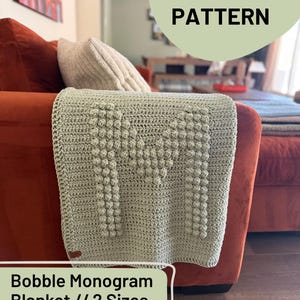 Può includere: Una coperta all'uncinetto verde salvia con un grande design a lettera "M" strutturato, drappeggiata su un divano color ruggine. La coperta presenta un motivo a punto nocciolina ed è etichettata come un modello all'uncinetto "Bobble Monogram Blanket".