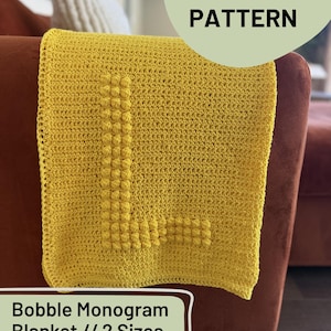 Könnte beinhalten: Eine gelbe Häkeldecke mit einem großen, strukturierten Buchstaben "L" in der Mitte. Die Decke ist über einem braunen Sessel drapiert. Der Text "Bobble Monogram Blanket // 2 Sizes" und "Crochet Pattern" ist sichtbar.