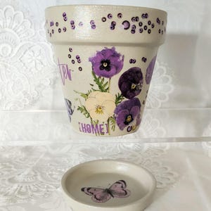 May include: A cream-colored ceramic flower pot and matching saucer featuring hand-painted purple pansies, scattered purple dots, and the text "HOME" in purple lettering. The pot has a glossy finish with a subtle shimmer and a butterfly design.