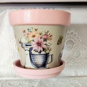 May include: A beige ceramic flower pot with a matching saucer, featuring a painted pink rim. The exterior displays a graphic of a stand mixer overflowing with colorful flowers, including pink, yellow, and white blossoms, along with small cupcake illustrations.
