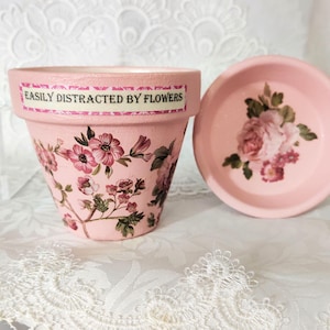 May include: A light pink flower pot and matching saucer featuring a floral pattern and the text EASILY DISTRACTED BY FLOWERS on the rim. The pot is decorated with pink and dark pink flowers with green leaves, while the saucer displays a rose motif.