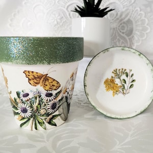 May include: A white ceramic plant pot with a glittery olive green rim, decorated with a yellow butterfly and various wildflowers. Beside it sits a matching white saucer featuring a small cluster of yellow flowers and a distressed green edge.