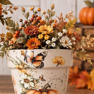 May include: A beige ceramic flower pot decorated with a painted jack-o'-lantern, monarch butterfly, and floral motifs, filled with an arrangement of artificial autumn flowers, orange berries, and green foliage.