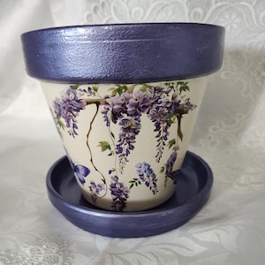 May include: A tapered ceramic flower pot with a matching saucer, both featuring a metallic dark purple rim and base. The cream-colored body is decorated with a detailed botanical print of hanging purple wisteria blossoms and green leaves.