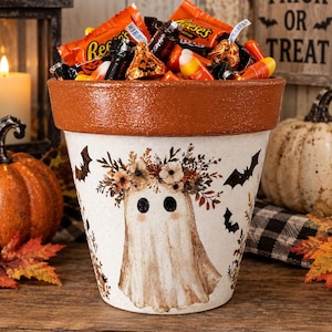 May include: A terracotta-colored clay pot featuring a white ghost illustration with a floral crown, surrounded by black bat silhouettes. The pot is filled with assorted Halloween candies, including Reese's cups, Hershey's Kisses, and candy corn.