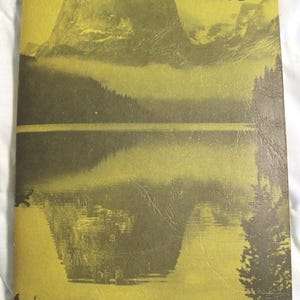 May include: A softcover book titled Wind River Trails by Finis Mitchell. The cover features a monochromatic yellow-green landscape illustration of a mountain reflected in a calm lake, with dark silhouettes of pine trees framing the bottom edges.