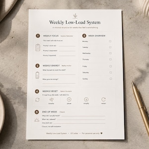 May include: A minimalist weekly planner titled "Weekly Low-Load System" with sections for focus, energy, and reset. The white paper has a simple layout with checkboxes and prompts. A gold pen and a ceramic mug are on the surface.