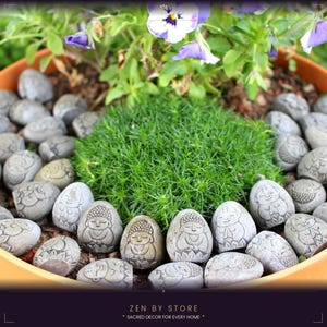 Pode incluir: Uma coleção de pequenas pedras cinzentas em forma de ovo com figuras gravadas de personagens sentados semelhantes a budas, dispostas num padrão circular em torno de um pedaço central de musgo verde num vaso laranja com flores roxas.