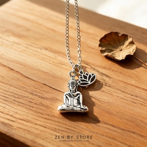 Könnte beinhalten: Eine silberfarbene Metallkette mit einem Anhänger in Form eines sitzenden Buddhas in Meditationshaltung und einem kleineren Lotusblüten-Anhänger. Die Kette besteht aus einfachen Gliedern. Der Text ZEN BY STORE SACRED DECOR FOR EVERY HOME ist unten aufgedruckt.