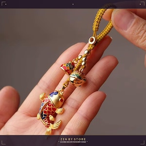 Peut inclure: Un charm en forme de poisson doré avec des détails en émail rouge, bleu et jaune, suspendu à un cordon tressé jaune. Le charm comprend deux petites cloches aux accents colorés. L'image est d'un objet décoratif.