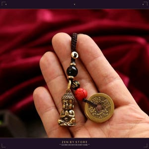 Puede incluir: Un amuleto de Buda de bronce con una moneda y cuentas negras y rojas. El amuleto está sujeto a un cordón negro. El texto "ZEN BY STORE" y "SACRED DECOR FOR EVERY HOME" está en la parte inferior de la imagen.