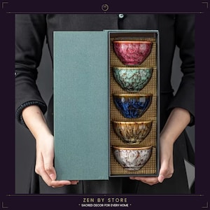 Könnte beinhalten: Ein Set aus fünf Keramikschalen in einer Geschenkbox. Die Schalen sind in den Farbtönen Rot, Grün, Blau, Braun und Weiß gehalten. Die Box ist türkis und die Schalen haben eine strukturierte, gesprenkelte Oberfläche. Der Text "ZEN BY STORE" und "SACRED DECOR FOR EVERY HOME" befindet sich unten.