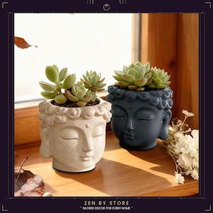 Puede incluir: Dos macetas de cerámica con forma de cabeza de Buda, una de color crema y otra gris oscuro, cada una con suculentas verdes. Las macetas están sobre una superficie de madera cerca de una ventana. El texto "ZEN BY STORE" está en la parte inferior.