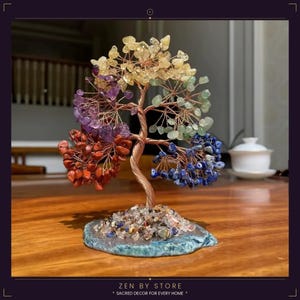 Könnte beinhalten: Eine dekorative Baumskulptur mit einem Stamm und Ästen aus Kupferdraht, verziert mit farbigen Edelsteinen, die die Chakren darstellen. Die Basis ist ein blau-grüner Stein, und der Baum steht auf einer Holzoberfläche. Der Text "ZEN BY STORE" ist enthalten.