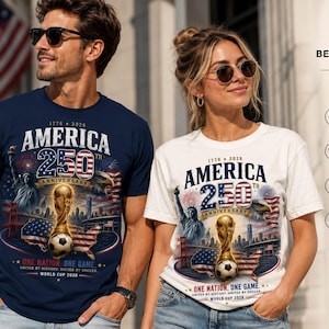 K&ouml;nnte beinhalten: Ein marineblaues T-Shirt und ein wei&szlig;es T-Shirt, beide mit einem Grafikdruck mit der Aufschrift America 250th Anniversary, 1776-2026, One Nation, One Game, World Cup 2026, mit einem Fu&szlig;ballpokal, einem Fu&szlig;ball, der Freiheitsstatue und einer US-Flagge.