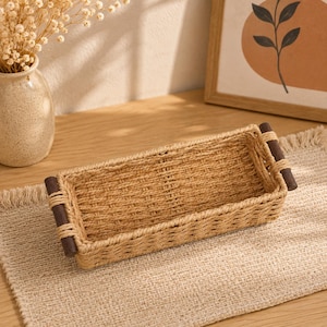 Peut inclure: Un panier rectangulaire tressé avec une texture en fibre naturelle beige clair et deux poignées cylindriques en bois brun foncé fixées aux extrémités. Le panier présente un motif tressé uniforme sur toute la base et les quatre parois latérales.