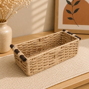 Peut inclure: Un panier rectangulaire tressé en fibre naturelle beige clair avec deux poignées en bois brun foncé fixées aux extrémités courtes. Le panier présente un dessus ouvert et un motif de tressage texturé sur les côtés et la base.