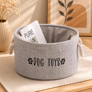 Peut inclure: Un panier de rangement en tissu gris avec des poignées en corde, portant l'inscription "DOG TOYS" en noir. Le panier contient un magazine avec le mot "PURE" visible. Conçu pour les fournitures pour animaux.